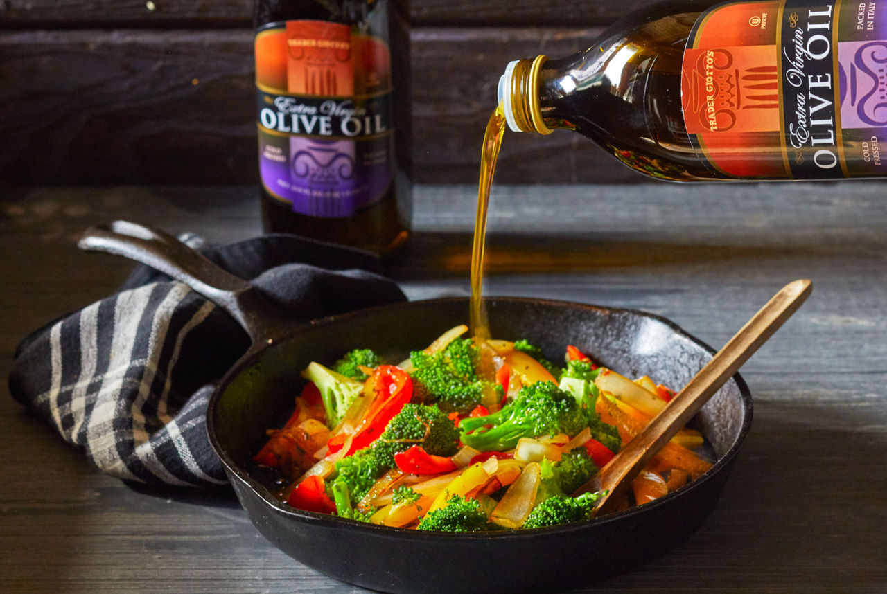 Trader Joe's Extra Virgin Olive Oil, being poured into cast iron skillet filled with colorful vegetables; dark, moody background