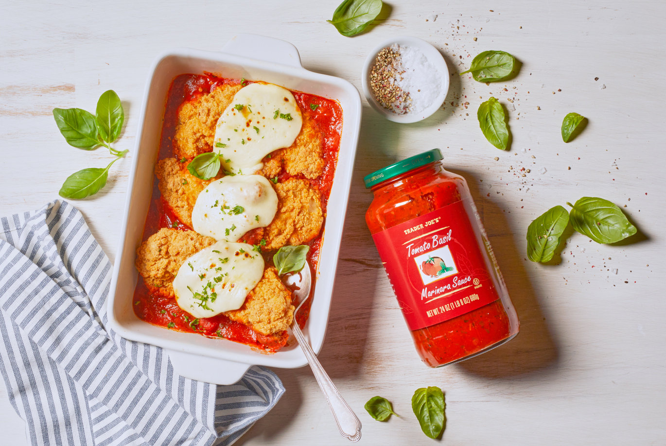Trader Joe's Tomato Basil Marinara Sauce; shown in a casserole pan with breaded chicken and mozzarella; basil leaves, pepper and salt on surface