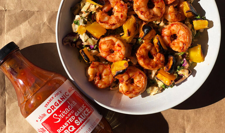 bowl of slaw with broiled spicy shrimp and charred mango chunks, next to a bottle of TJ's Organic Sriracha & Roasted Garlic BBQ Sauce
