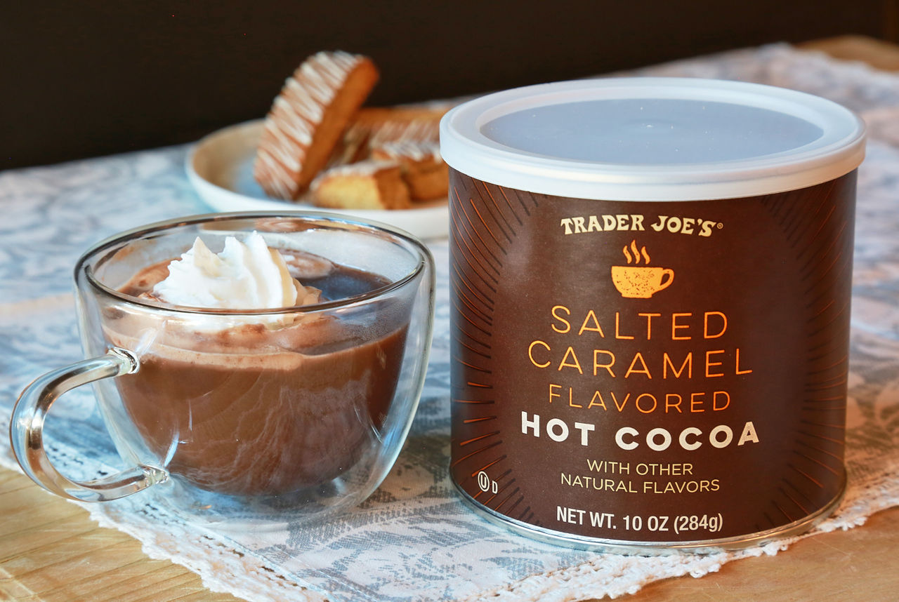 Trader Joe's Salted Caramel Hot Cocoa; in a glass mug topped with whipped cream, plate of biscotti cookies in the background