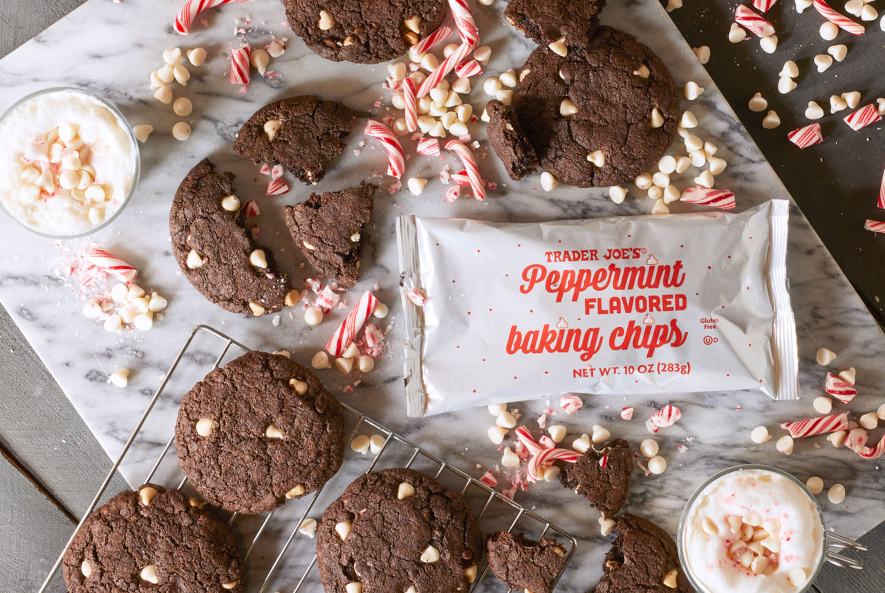 Trader Joe's Peppermint Flavored Baking Chips, shown baked into chocolate peppermint cookies; more chips and broken pieces of peppermint candy cane scattered on surface. Two mugs of hot white, frothy beverage topped with chips