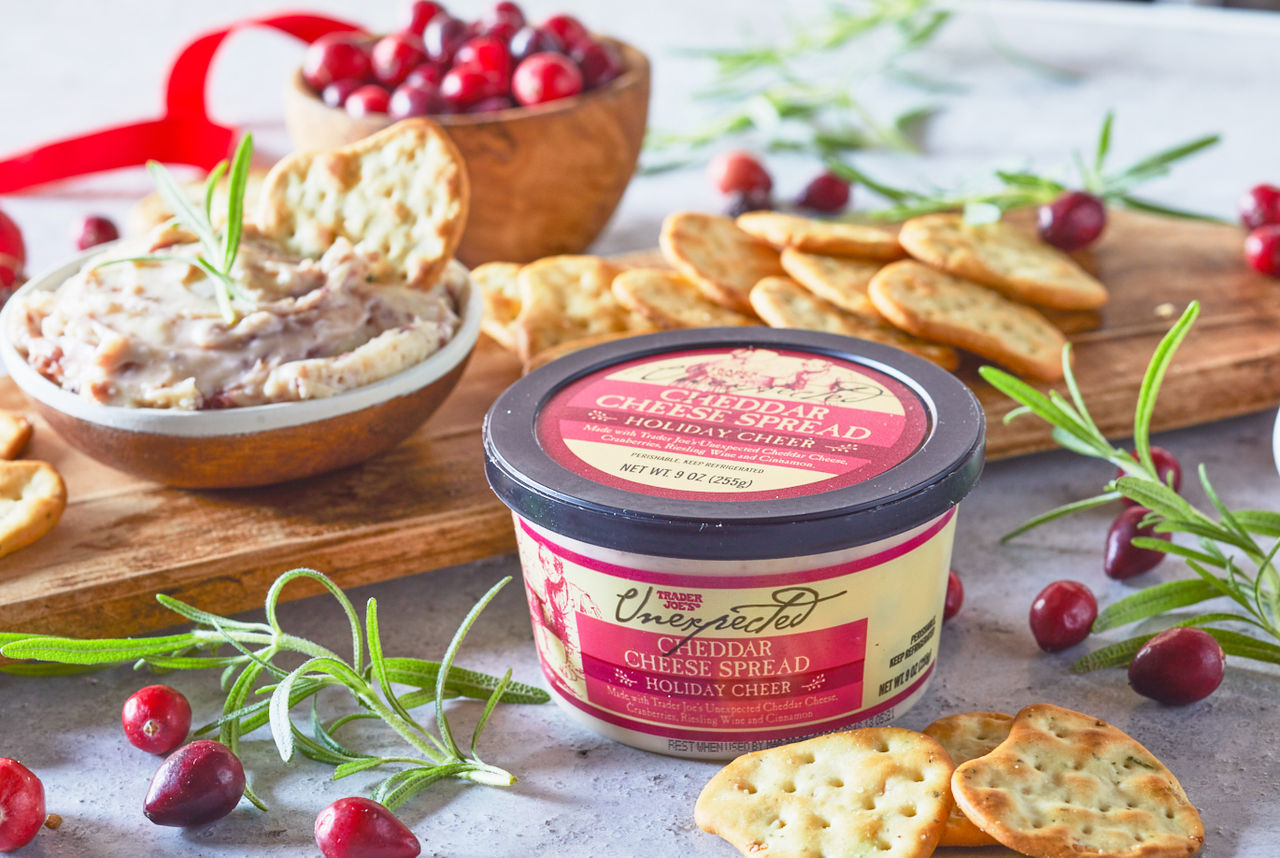 Trader Joe's Holiday Cheer Unexpected Cheddar Cheese Spread; in small bowl surrounding by Pita Bite Crackers, garnished with fresh rosemary and cranberries; festive ribbon and ornaments in background