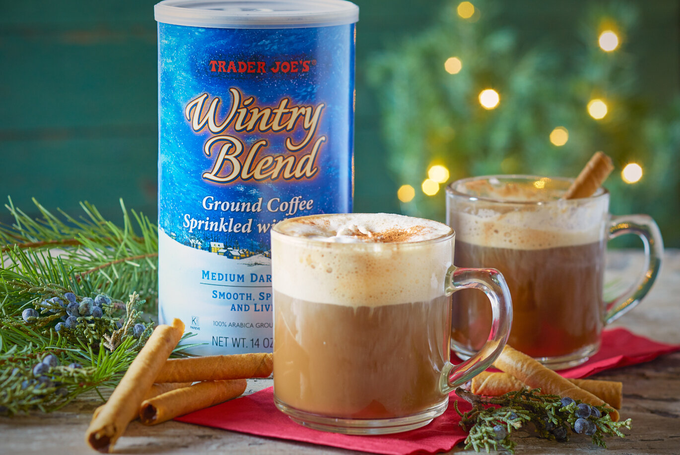 Trader Joe's Wintry Blend Ground Coffee prepared in two glass mugs, topped with frothy cream and cinnamon; a festive background with pine branches and lights